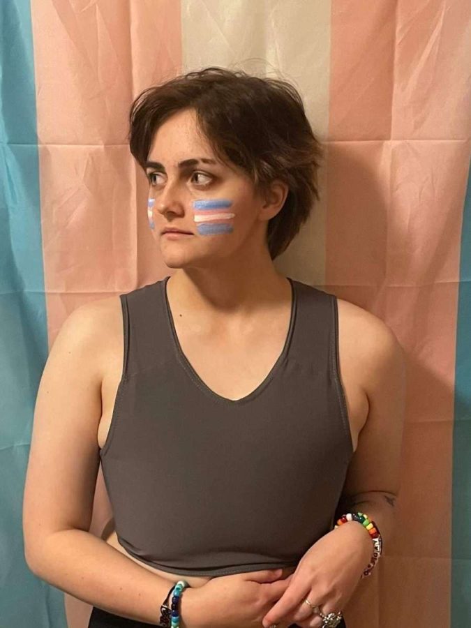 UNC Asheville senior Kai Tilly dawning face paint, standing in front of the Trans flag.