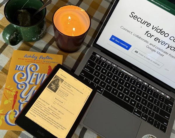 On the table sits a laptop with Google Meet pulled up on it, a candle burning, "The Seven Year Slip" by Ashley Poston in paperback, a kindle and a cup of tea. 